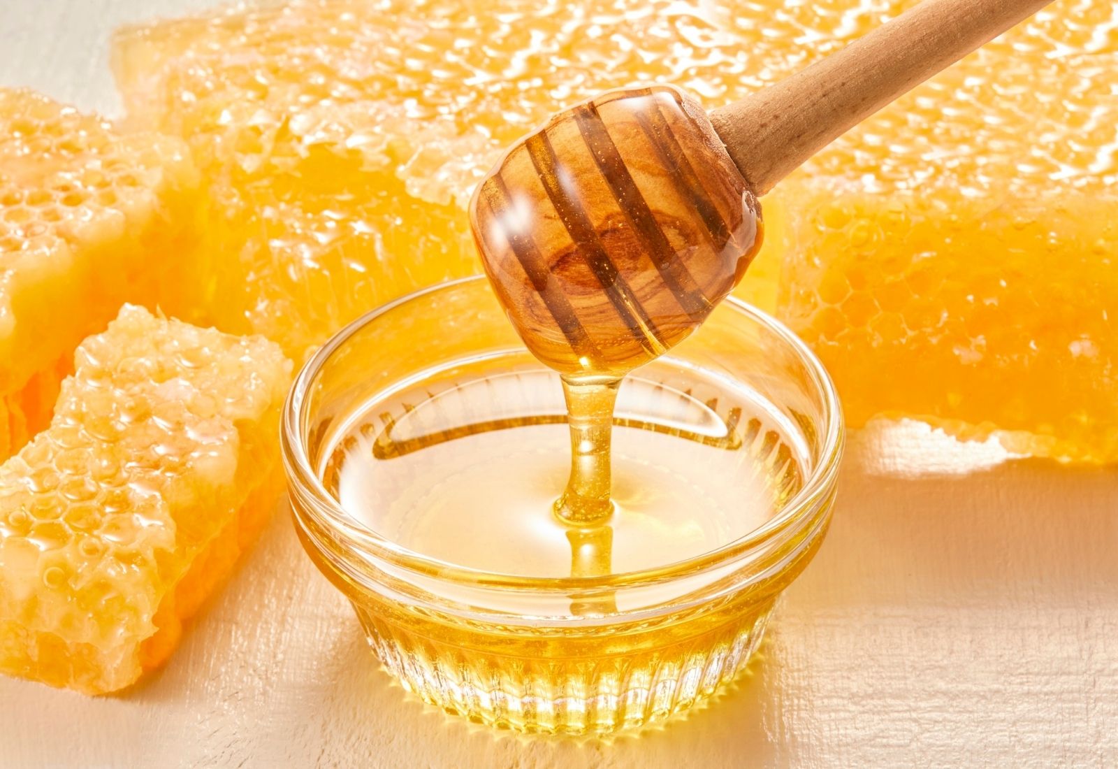Golden honey flowing from a wooden dipper into a glass bowl, with fresh honeycomb pieces in the background.