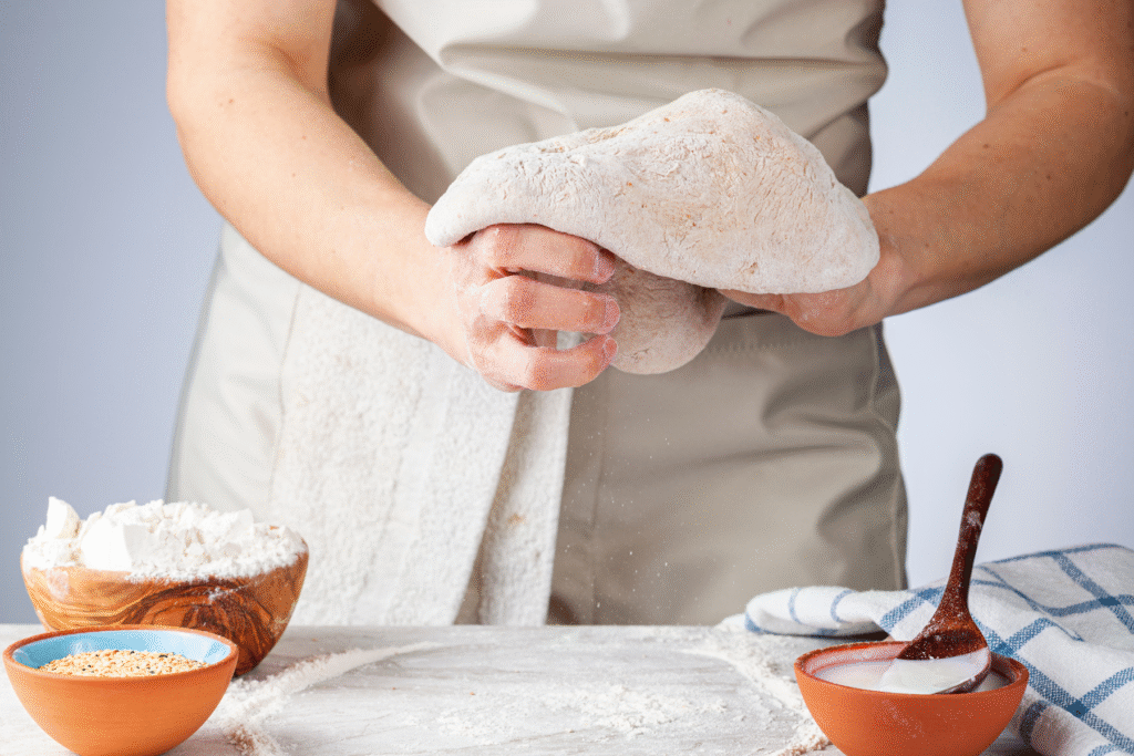 baker shaping and working sourdough dough by hand on a floured counter with bowls of flour and seeds nearby